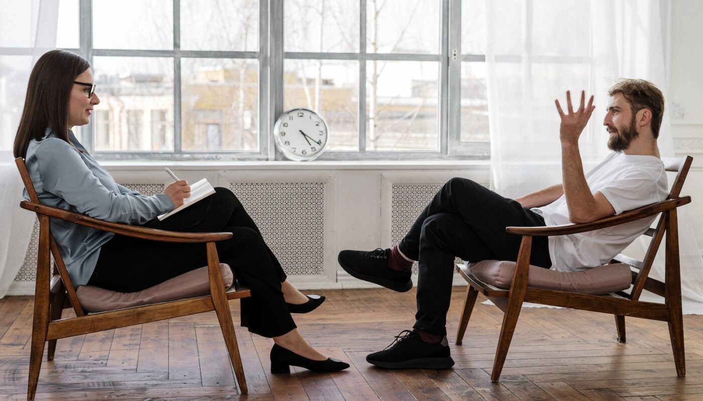 Person in Black Pants and Black Shoes Sitting on Brown Wooden Chair