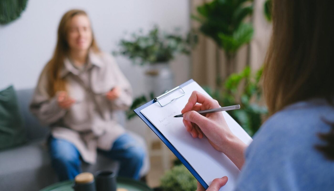 Unrecognizable professional female psychologist writing on clipboard while sitting against client on blurred background during psychotherapy session in light office