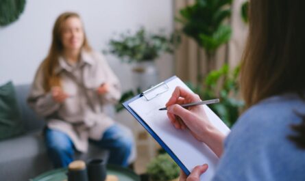 Unrecognizable professional female psychologist writing on clipboard while sitting against client on blurred background during psychotherapy session in light office